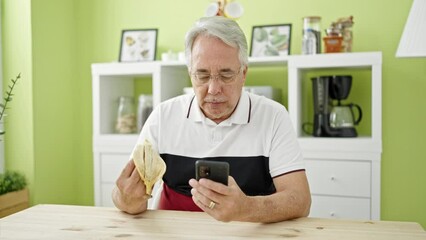 Wall Mural - Middle age man with grey hair sitting eating banana using smartphone at dinning room