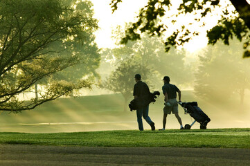 Two men golfers walking on a golf course with the sun streaking through the trees on a beautiful summer morning