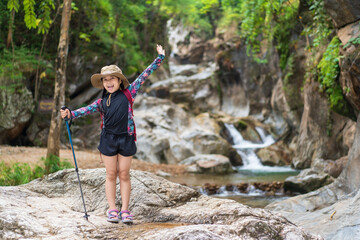 Wall Mural - asian child hiking or kid girl happy carry backpack with walking pole or people smile trekking camping to standing on rocks and nature waterfall in forest for holiday travel adventure in national park