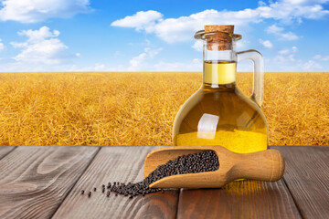 Wall Mural - rapeseed oil in bottle with rape seeds in scoop on table and canola field as background