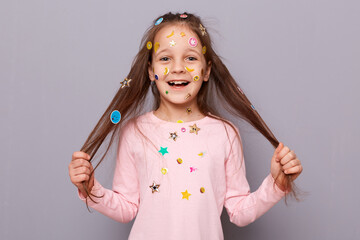 Excited amazed smiling little girl covered colorful kid's stickers standing isolated over gray background pulling her hair aside being joyful and pleasantly surprised.