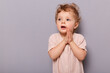 © sementsova321 - Portrait of amazed little caucasian infant girl with blond wavy posing isolated over gray background keeps hand together looking away at copy space for promotion.