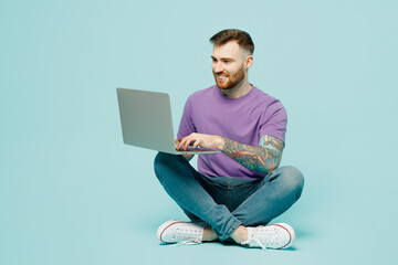 Full body happy fun programmer young IT man he wears purple t-shirt sitting hold use work on laptop pc computer isolated on plain pastel light blue cyan background studio portrait. Lifestyle concept.