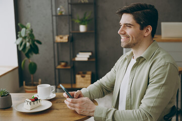 Wall Mural - Side viw young minded happy smiling man wears casual clothes sits alone at table in coffee shop cafe restaurant indoors work hold use mobile cell phone look aside rest relax during free time inside.
