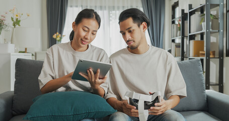 Poster - Young Asian couple sit on couch hold digital tablet and wear virtual reality glasses are watching and show imagine spend time together have fun at home on weekend. Lifestyle tech innovation concept.