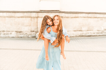 Mother of the daughter walks playing. Mother holds the girl on her back, holding her legs, and her daughter hugs her by the shoulders. Dressed in blue dresses.