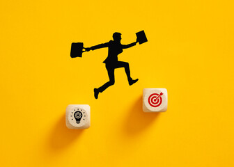Silhouette of a businessman jumping from idea to goal symbol on wooden cubes.