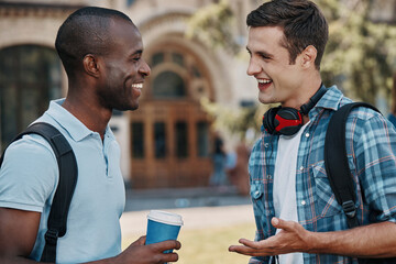 Two young men talking and smiling while standing against University building together