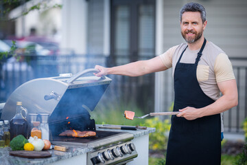 Wall Mural - Male chef grilling and barbequing in garden. Barbecue outdoor garden party. Handsome man preparing barbecue meat. Concept of eating and cooking outdoor during summer time.