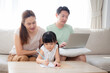 © NT_studio - Happy family with mother teaching homework with daughter and father working with laptop on sofa in living room at home, mom explaining schoolwork with kid together, lifestyles and education.