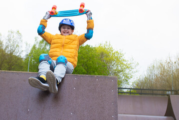 Wall Mural - portrait of a smiling schoolboy in a safety helmet with a bright blue skateboard, an urban cruiser in his hands in an outdoor skate park in spring, autumn