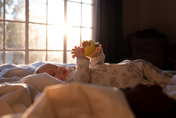 Wall Mural - A curious, happy and content baby lying on a bed, playing with a yellow toy in the early morning sunlight