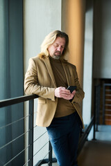 Poster - Elegant bearded mature businessman in formalwear texting in mobile phone while standing by railings inside modern office building