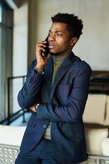 Poster - Young serious African American businessman in dark blue suit standing in front of camera and speaking to colleague on smartphone
