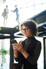 Poster - Young smiling female manager in eyeglasses and smart casualwear watching video in smartphone while standing against two men