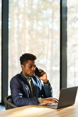 Poster - Young serious African American broker speaking on smartphone and typing while sitting by workplace in front of laptop against window
