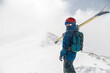 man stands with his back to the camera with alpine skis on his shoulder against the backdrop of a mountain during a snowfall