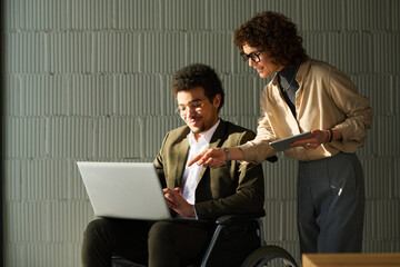 Poster - Young businesswoman with tablet bending over male colleague in wheelchair and pointing at screen of laptop on his knees
