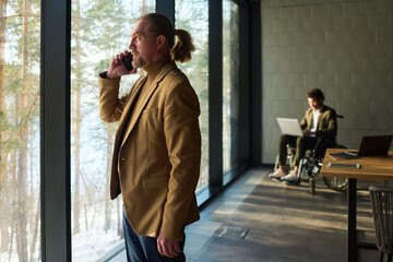 Poster - Confident mature ceo in beige quiet luxury attire talking on smartphone while standing in front of large window against coworker in wheelchair