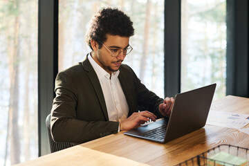 Poster - Serious young analyst or broker looking at laptop screen by workplace while analyzing online financial data or working over new project