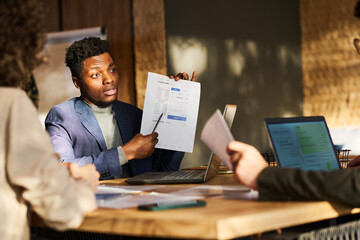 Poster - Young confident AFrican American male broker pointing at document with financial data and explaining it to colleagues at meeting in boardroom