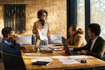 Wall Mural - Confident business coach pointing at financial document with graphic data while explaining it to group of multicultural coworkers at meeting