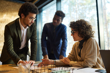 Confident businesswoman explaining financial data to young intercultural male colleagues standing in front of her at meeting in boardroom