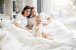 © sofiko14 - Cheerful little kid in white baby clothes resting on bed while joyful adult people staying under blanket in bright bedroom interior. Loving mother and father telling nursery rhyme from verse book.