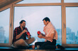 © Anna - Young men in home clothes are having dinner near the window of the house. A couple of men in love are sitting on the windowsill
