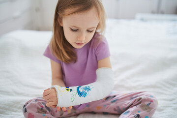 Adorable preschooler girl with a broken arm at home on the bed draws with felt-tip pens on an orthopedic cast