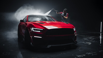 Adult Car Washer in Uniform Washing a Red Performance Car with a High Pressure Cleaner. Cleaning Technician Working on a Stylish American Car in a Dark Room. Commercial Studio Footage for Advertising