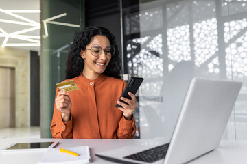 Cheerful and successful hispanic businesswoman working inside office, woman holding bank credit card and phone, online shopping and successful booking, woman at workplace with laptop.