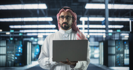 Wall Mural - Portrait of a Muslim Data Center Technical Manager Wearing Traditional White Clothes, Using Laptop Computer in a Modern Technological Server Farm Facility. Arab Cybersecurity Expert at Work