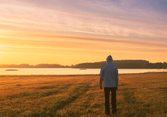 Wall Mural - Young caucasian man in hoodie walking to pond under dramatic sunrise sky. Czech landscape