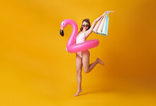 Happy Sexy Young Woman Dressed In Swimwear Holding Shopping Bag And Flamingo Rubber Ring Jumping For Summer Sale On Yellow Background.