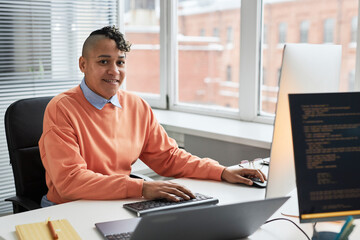Portrait of young programmer smiling at camera while working with security codes on computer sitting at his workplace