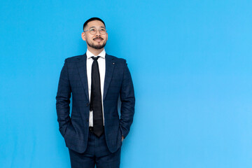 Wall Mural - successful asian businessman in suit stands on blue isolated background and smiles, korean entrepreneur in glasses