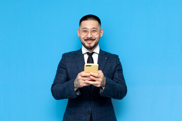 Wall Mural - successful asian businessman in suit use smartphone and smile on blue isolated background, korean entrepreneur