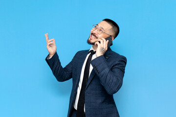 Wall Mural - successful asian businessman in suit and glasses is talking on the phone on blue isolated background