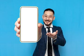 successful asian businessman in suit and glasses shows blank smartphone screen on blue isolated background