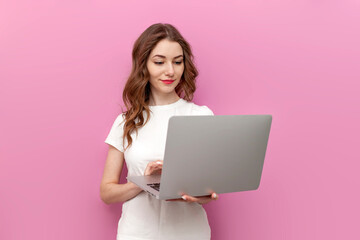 young cute woman in white t-shirt uses laptop on pink isolated background, woman freelancer types on
