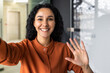 Beautiful business woman with curly hair wearing looking at smartphone camera, waving her hand in greeting gesture, talking with friends and colleagues, using video call app.
