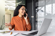 © Liubomir - Young successful hispanic businesswoman working inside modern office, female employee cheerfully talking to colleagues on phone and smiling, using laptop while sitting at desk.