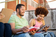 Two young, dark-skinned millennials sit on some outdoor steps. The chubby Dominican boy is writing in a book while the girl with the afro hair is studying on a tablet. Concept of new technologies.