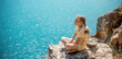 © svetograph - Happy girl perched atop a high rock above the sea, wearing a yellow jumpsuit and braided hair, signifying the concept of summer vacation at the beach.