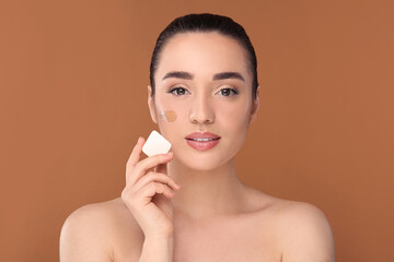 Wall Mural - Woman applying foundation on face with makeup sponge against brown background