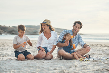 We never stop laughing around our children. Full length shot of a happy couple sitting with their two children who are blowing bubbles on the beach.