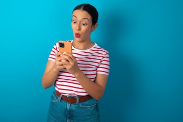 Wall Mural - beautiful woman wearing striped T-shirt over blue studio background looks with bugged eyes, holds modern smart phone, receives unexpected message from friend, reads reminder.