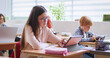 © ihorvsn - Caucasian schoolgirl in glasses sitting at desk in classroom, writing and using tablet device for study. Time at school. Smart girl learning at lesson and checking exercise in copybook with computer.