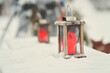 Rustic wooden lanterns on tables outside, red candles inside the lanterns. Tables are covered in snow and lanterns are sitting on top of the snow surface, one lantern in focus. Snowy winter day.